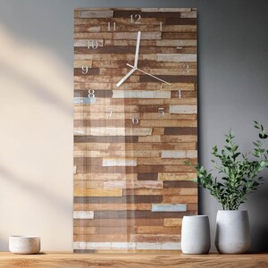 Op de afbeelding: Een rechthoekige wandklok met een houten plankontwerp. De wijzerplaat heeft zilveren cijfers en witte wijzers. De klok wordt weergegeven op een houten oppervlak, met twee witte vazen en een kleine kom. De achtergrond is een neutraal grijs.