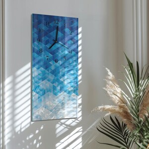 May include: A rectangular wall clock with a geometric design in shades of blue. The clock face features black hands and numerals. The background has a pattern of blue triangles. The clock is mounted on a white wall.