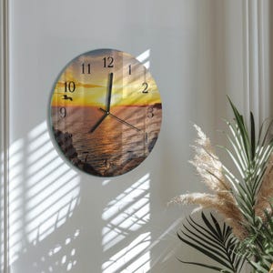 May include: A round wall clock featuring a sunset over the ocean with rocky cliffs in the foreground. The clock face displays black numbers from 1 to 12 and black hour, minute, and second hands. A small silhouette of a bird is visible on the left.