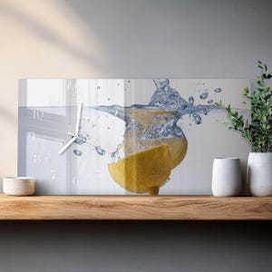 May include: A rectangular wall clock with a lemon slice submerged in water. The clock face has white numerals and hands. The image features a splash of water and a bright yellow lemon, creating a refreshing and vibrant aesthetic. The clock is displayed on a wooden shelf.