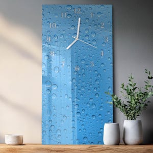 May include: A rectangular wall clock with a blue background featuring a water droplet design. The clock has gold-colored numerals and hands. The clock is displayed on a wooden surface with decorative white vases and a plant.