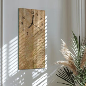May include: A rectangular wall clock with a natural leaf pattern. The clock face features black numerals and hands. The clock is mounted on a white wall, with shadows from a window and plants in the background.