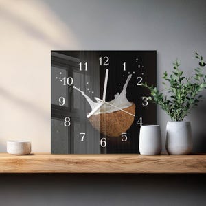 May include: A square wall clock featuring a black background with a central image of a halved coconut splashing with white liquid. The clock face displays white Arabic numerals from 1 to 12 and two thin white clock hands centered on the coconut.