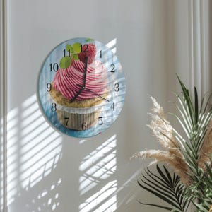 May include: Round wall clock featuring a cupcake design. The clock face displays a cupcake with pink frosting, a raspberry, and green leaves. The clock has black hands and a blue background. The numbers are in black.