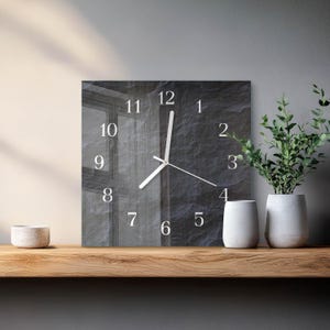 Könnte beinhalten: Eine quadratische Wanduhr mit einem dunkelgrauen, strukturierten, steinartigen Hintergrund und weißen arabischen Ziffern von 1 bis 12, die kreisförmig angeordnet sind. Die Uhr hat drei dünne weiße Zeiger, die die Zeit auf der dunklen Fläche anzeigen.