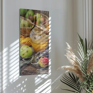 Op de afbeelding: Een rechthoekige wandklok met een stilleven van appels, honing en brood. De klok heeft een afbeelding van een pot honing met een honingdipper, appels en een broodmand. De wijzers van de klok staan op 10:08.