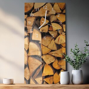 May include: A rectangular wall clock with a woodpile design. The clock face has white numerals and hands, set against a backdrop of stacked, cut logs in shades of brown and tan. The clock is mounted on a grey wall, with decorative vases and plants on a wooden surface.
