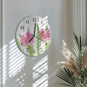 Könnte beinhalten: Eine runde Wanduhr mit einem Blumendesign aus rosa Tulpen und grünen Blättern. Die Uhr hat schwarze Zeiger und Ziffern, mit den Zahlen 1 bis 12. Die Uhr ist an einer weißen Wand befestigt.