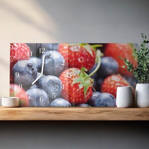 May include: Rectangular wall clock featuring a vibrant image of fresh strawberries and blueberries. The clock face displays black numerals and hands against the fruit background. The clock is mounted on a wooden shelf.