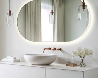 Puede incluir: Un tocador de baño ovalado blanco con un lavabo de cerámica blanca, un grifo de cobre y tres pomos de cobre. El tocador tiene dos cajones y está cubierto con una encimera blanca. Un espejo ovalado blanco con una luz LED integrada está montado sobre el lavabo. Dos lámparas colgantes de vidrio transparente con detalles de cobre cuelgan del techo. Un jarrón con flores blancas se encuentra sobre la encimera.