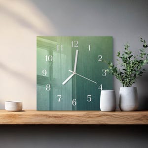 May include: Square wall clock with a gradient green and blue background. The clock face features white numerals and hands. The clock is displayed on a wooden shelf with decorative white vases and a potted plant.