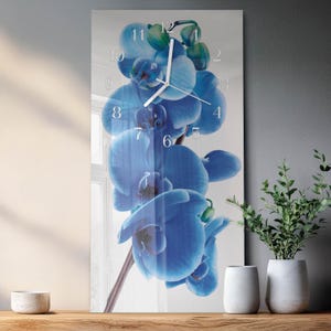 Può includere: Un orologio da parete rettangolare con un vibrante design di orchidea blu. Il quadrante dell'orologio mostra numeri e lancette bianche, sullo sfondo del fiore. L'orologio è esposto su una superficie di legno, con vasi bianchi e verde sullo sfondo.