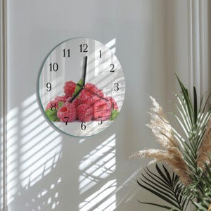 May include: A round wall clock with a glass face. The clock face features a vibrant image of fresh raspberries. Black numerals mark the hours, and black hands indicate the time. The clock is mounted on a white wall.