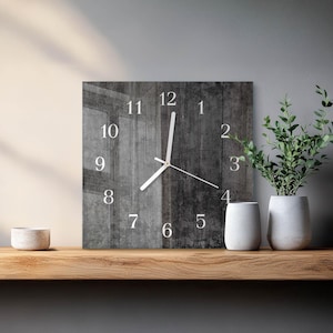 May include: Square wall clock with a grey wood grain design. The clock face features white numerals and hands. The clock is displayed on a wooden shelf with decorative white vases and a potted plant.