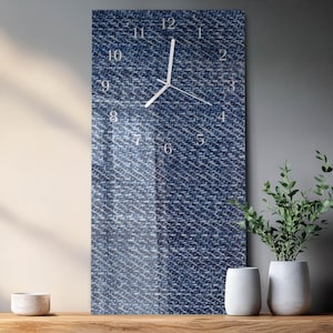 May include: A rectangular wall clock with a denim texture background. The clock face features white numerals and hands. The clock is mounted on a wall, with a wooden surface and decorative plants in the foreground.