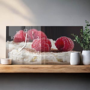 May include: A rectangular wall clock featuring a close-up of a dessert with fresh raspberries. The clock face displays white numbers and hands. The dessert has a white creamy base, topped with raspberries and chocolate shavings.