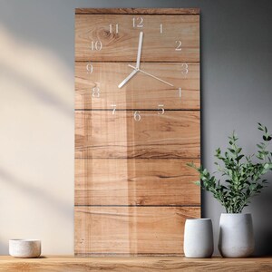 May include: A rectangular wall clock with a wooden plank design. The clock face features white numerals and hands. The clock is mounted on a wall with a neutral color scheme. The clock is approximately 12 inches wide and 24 inches tall.