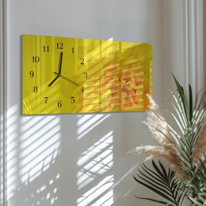 May include: A rectangular wall clock with a bright yellow face. Black numerals mark the hours, and black hands indicate the time. The background features a blurred floral design. The clock is mounted on a white wall.