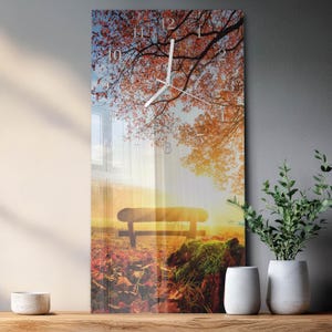Könnte beinhalten: Eine rechteckige Wanduhr mit einer herbstlichen Landschaft mit einer Bank, Bäumen und einem Sonnenuntergang. Das Zifferblatt hat weiße Zeiger und Ziffern. Das Bild wird von einer Holzoberfläche und einer grauen Wand umrahmt, mit dekorativen Pflanzen in weißen Töpfen.