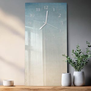 May include: A rectangular wall clock with a gradient blue and beige background, featuring a window and sheer curtains. The clock has white hands and numerals. Two white vases and a potted plant are on a wooden surface.