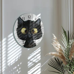 Könnte beinhalten: Eine runde Wanduhr mit einer Illustration einer schwarzen Katze mit großen gelben Augen. Das Zifferblatt zeigt schwarze Ziffern auf weißem Hintergrund. Die Uhr ist an einer weißen Wand befestigt. Durchmesser ca. 25 cm.