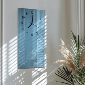 May include: A rectangular wall clock with a blue textured background. The clock has black numerals and hands. The clock is mounted on a white wall, with shadows from a window visible on the wall and clock.