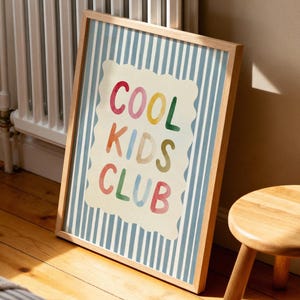 May include: A framed print with the words "COOL KIDS CLUB" in colorful letters. The background has blue and white vertical stripes. A small wooden stool sits nearby on a wooden floor.