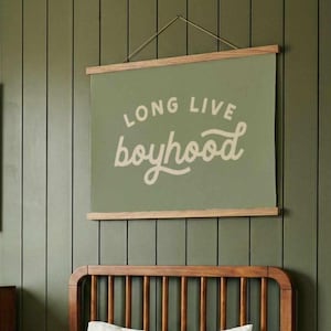 May include: A room with a green wall, a wooden bed, and a framed landscape painting. A green poster with the words "LONG LIVE boyhood" hangs above the bed. Two pillows with a longhorn design rest on the bed.