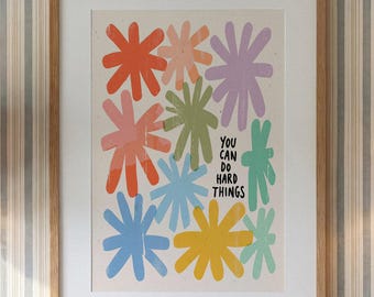 May include: A framed wall art print featuring a cream background with various hand-drawn style starburst shapes in orange, pink, light green, lavender, blue, yellow, and teal. The text "YOU CAN DO HARD THINGS" is printed in black.