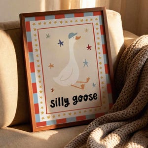 May include: A framed print featuring a whimsical illustration of a white goose wearing a blue cap, surrounded by stars. The words "silly goose" are written in black script below the goose. The frame has a brown wood finish with a red, blue, and gold border.