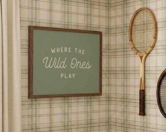 May include: A framed sign with the text "WHERE THE Wild Ones PLAY" hangs on a wall with plaid wallpaper. Two vintage wooden tennis rackets are mounted on the wall. The room has a classic, vintage aesthetic.