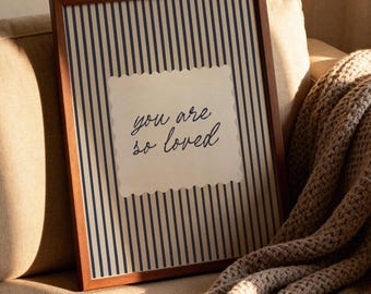 May include: A rectangular wall art piece in a dark wood frame featuring a cream-colored background with vertical navy blue stripes. In the center, a cream-colored scalloped rectangle displays the dark blue cursive text, you are so loved.