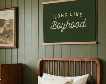 May include: A rectangular wall hanging featuring a dark green fabric banner with the cream-colored text "LONG LIVE Boyhood" printed in a mix of block and script fonts, suspended from a light-colored wooden frame with a thin hanging string.
