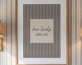 May include: A rectangular wall art print in a light wood frame featuring a vertical navy blue and beige striped background with a central scalloped-edge beige square containing the dark blue cursive text "how lucky are we".