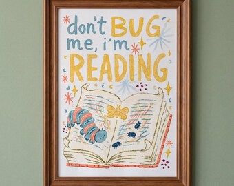 May include: A framed print featuring an open book with a blue and red striped caterpillar, a yellow moth, and small dark beetles. The text reads, "don't BUG me, i'm READING" in blue and yellow letters, surrounded by colorful star and dot accents.
