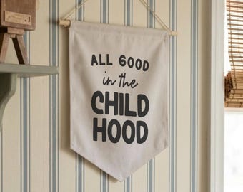 May include: A cream-colored fabric wall banner hanging from a wooden dowel with a string. The banner features the black printed text ALL GOOD in the CHILD HOOD. The banner is displayed against a wall with vertical blue and white stripes.