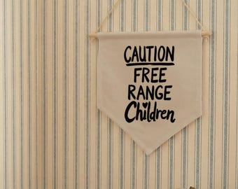 May include: A beige canvas wall banner featuring the black text CAUTION FREE RANGE Children, suspended by a wooden dowel and string. The banner is displayed on a wall with vertical blue and white stripes, near a brown leather satchel.