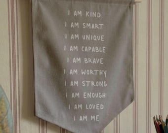 May include: A grey fabric wall banner hanging from a wooden dowel with white text that reads: I AM KIND, I AM SMART, I AM UNIQUE, I AM CAPABLE, I AM BRAVE, I AM WORTHY, I AM STRONG, I AM ENOUGH, I AM LOVED, I AM ME. The banner has a pointed bottom edge.