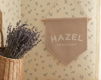 May include: A woven wicker basket filled with lavender, hanging on a wooden hook. A pink pennant with the name "HAZEL PENELOPE" hangs on the wall. The background is floral wallpaper.