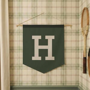 May include: A dark green pennant with a large white letter "H" hangs on a wall with plaid wallpaper. Two vintage tennis rackets are mounted on the wall. A cream-colored curtain is on the left.