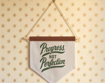 May include: A white fabric wall banner featuring the dark green text Progress NOT Perfection. The banner is suspended from a wooden dowel with a tan rope and a metal hook, featuring a brown top border and a pointed bottom edge.