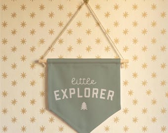 May include: A sage green banner with the words "little EXPLORER" in white script and block letters, with a small tree graphic. The banner is suspended by a wooden dowel and natural rope, hanging against a cream-colored wall with gold star patterns.