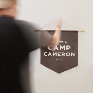Peut inclure: Une banni&egrave;re murale en tissu marron fonc&eacute; avec le texte blanc WELCOME TO CAMP CAMERON EST 2024 et deux petites ic&ocirc;nes de sapins verts. La banni&egrave;re est suspendue &agrave; une fine tige en bois par une ficelle claire sur un mur blanc uni.