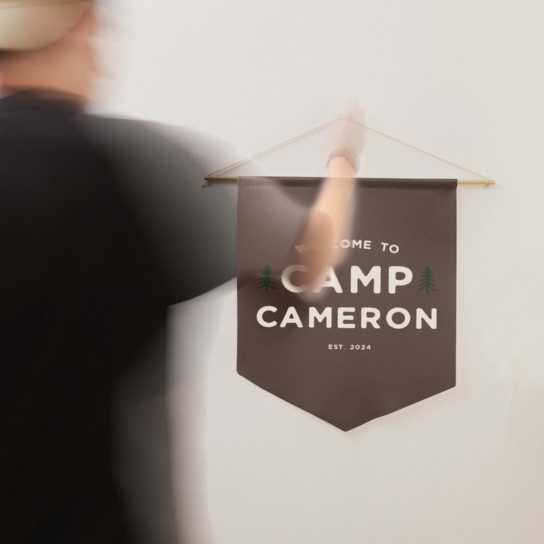 Peut inclure: Une banni&egrave;re murale en tissu marron fonc&eacute; avec le texte blanc WELCOME TO CAMP CAMERON EST 2024 et deux petites ic&ocirc;nes de sapins verts. La banni&egrave;re est suspendue &agrave; une fine tige en bois par une ficelle claire sur un mur blanc uni.