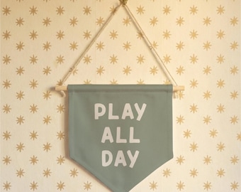 May include: A sage green banner with the words "PLAY ALL DAY" in white block letters. The banner is suspended from a wooden dowel and rope, hanging against a cream-colored wall with gold star patterns.