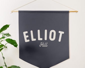 May include: A dark blue fabric banner with white text that reads "ELLIOT Hill". The banner is hanging from a wooden dowel rod with white string.