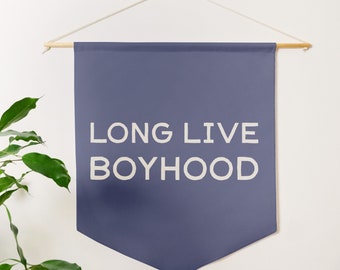 May include: A blue banner with white text that reads "LONG LIVE BOYHOOD". The banner is hanging from a wooden dowel rod with white string.