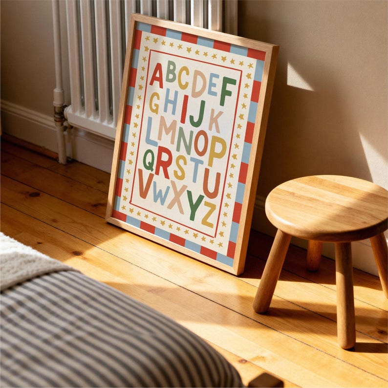 May include: A framed alphabet print with a wooden frame. The print features the letters A through Z in various colors, with a red and blue striped border and gold stars. The print is leaning against a wall on a wooden floor.
