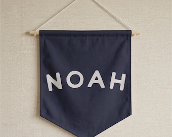May include: A dark navy blue fabric wall banner featuring the name NOAH printed in large, bold, white capital letters. The banner is suspended from a light-colored wooden dowel rod by a thin, white cord attached to both ends of the rod.