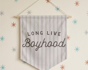 May include: A fabric wall banner featuring a light gray and white vertical pinstripe pattern. The text LONG LIVE Boyhood is printed in dark gray lettering across the center. The banner hangs from a wooden dowel with a thin white string.
